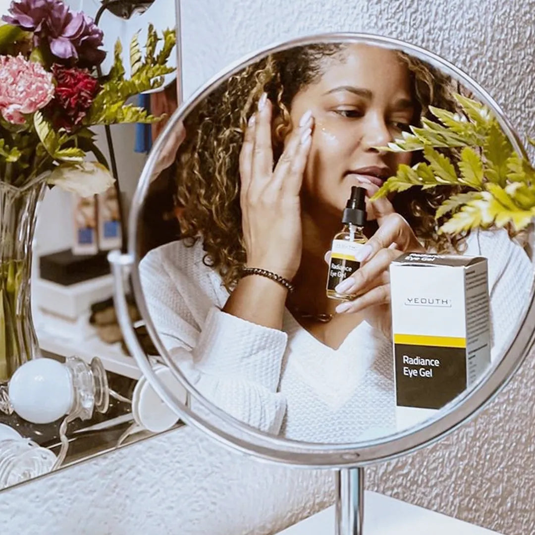 Woman applying YEOUTH Radiance Eye Gel in front of mirror with product bottle and box visible on vanity.