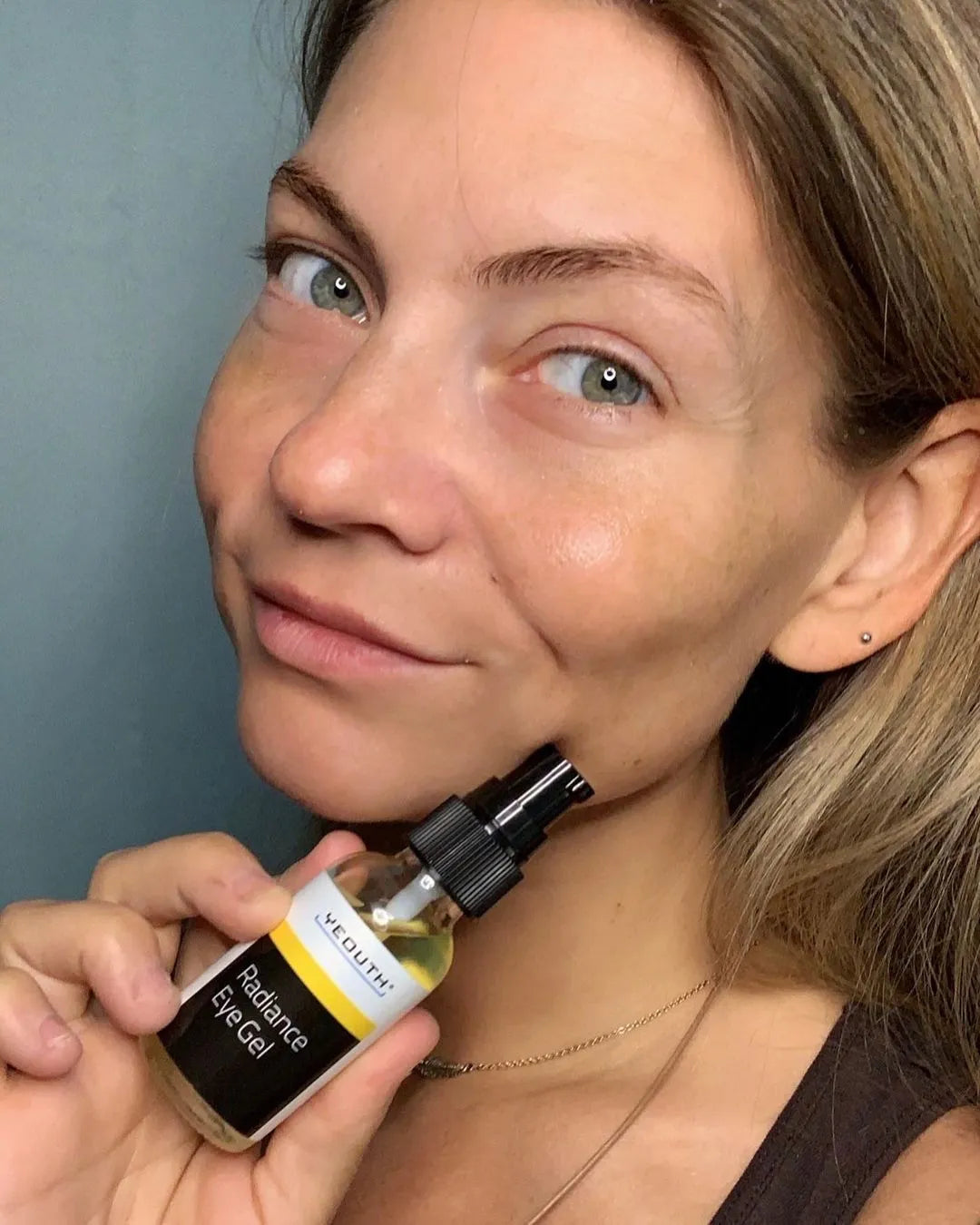Woman holding YEOUTH Radiance Eye Gel close to her face, showing natural skin and clean skincare product use.