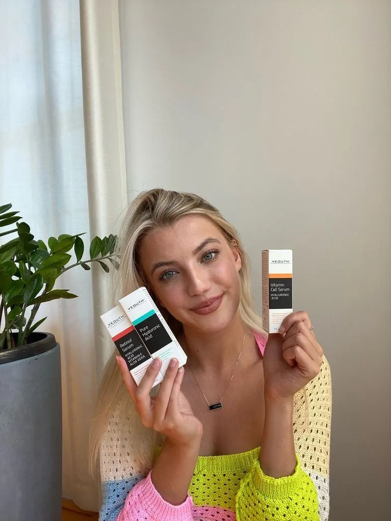 Face Serum Set Woman smiling while holding YEOUTH Vitamin C&amp;E Serum and Pure Hyaluronic Acid, seated indoors beside a plant.